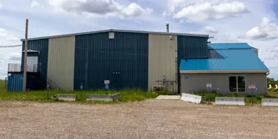 Exterior view of the Clearview Arena in Goodlow, British Columbia. The building features grey horizontal siding, a bright blue metal roof, and several large windows under a partly cloudy sky.