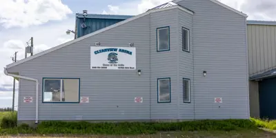 Exterior view of the Clearview Arena in Goodlow, British Columbia. The building features grey horizontal siding, a bright blue metal roof, and several large windows under a partly cloudy sky.