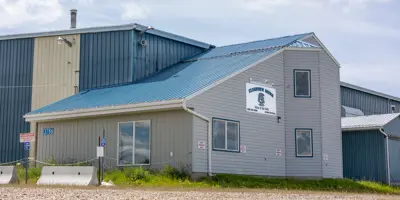 Exterior view of the Clearview Arena in Goodlow, British Columbia. The building features grey horizontal siding, a bright blue metal roof, and several large windows under a partly cloudy sky.